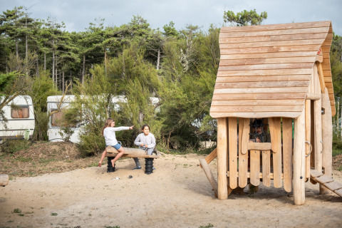 Dos niños juegan en un balancín junto a una casita de madera en un parque de vacaciones rodeado de árboles y caravanas.