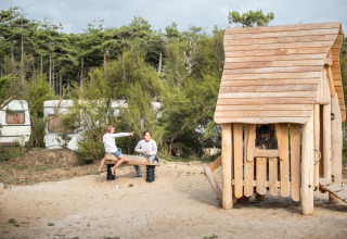 Twee kinderen spelen op een wip aan een houten speelhuisje in een vakantiepark omringd door bos en caravans.