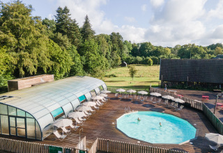 Udendørs swimmingpool med dæk, liggestole og parasoller på Huttopia Calvados - Normandie i en grøn skovrig region.
