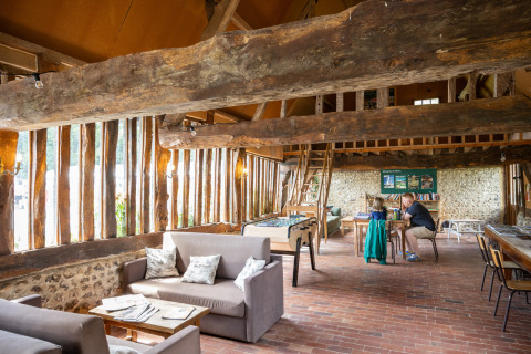 Intérieur convivial à Huttopia Calvados - Normandie, poutres en bois, canapés, jeux de société et ambiance chaleureuse.