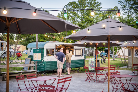 Zona de cafetería al aire libre con luces y sombrillas junto a una food truck vintage en Huttopia Calvados, Normandía.