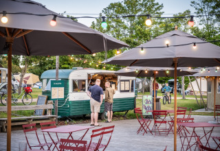 Udendørs caféområde med parasoller og lyskæder ved en vintage madvogn i Huttopia Calvados, Normandiet.