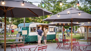 Zona de cafetería al aire libre con luces y sombrillas junto a una food truck vintage en Huttopia Calvados, Normandía.