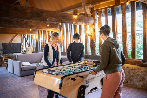 Drie personen spelen tafelvoetbal in een warme, landelijke hut met houten balken bij Huttopia Calvados.