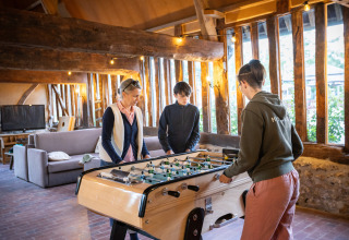 Drie personen spelen tafelvoetbal in een warme, landelijke hut met houten balken bij Huttopia Calvados.