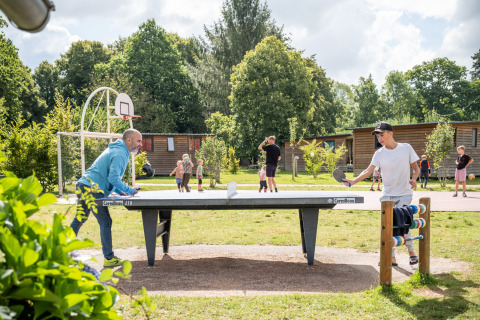 Personas juegan al ping pong al aire libre en Huttopia Calvados - Normandie, rodeados de naturaleza y familias.