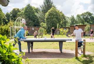 Folk spiller bordtennis udendørs på Huttopia Calvados - Normandie, med børn og grønne omgivelser i baggrunden.