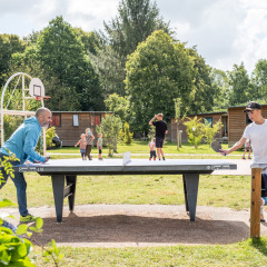 Personas juegan al ping pong al aire libre en Huttopia Calvados - Normandie, rodeados de naturaleza y familias.