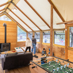 Interior acogedor en Huttopia Lac de Sillé, Francia, con familias jugando y grandes ventanas al bosque.