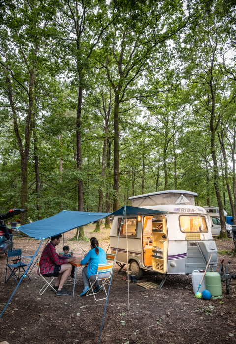 Gezin kampeert bij Huttopia Lac de Sillé, Pays de la Loire, Frankrijk, met caravan en luifel onder de bomen.