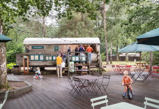 Børnevenligt udendørs caféområde ved Huttopia Lac de Sillé, med træmøbler og grønne omgivelser.