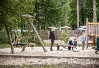 Børn leger og en voksen overvåger i en legeplads med sand og gynger ved Huttopia Lac de Sillé, Frankrig.