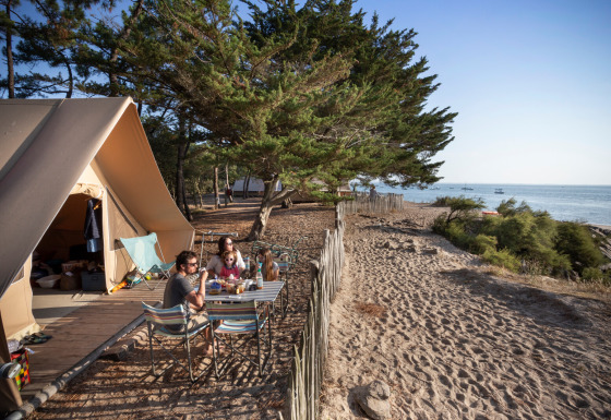 Famiglia che mangia davanti a una tenda safari Toile&Bois sulla costa, con vista sul mare e alberi attorno.