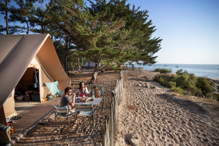 Famille prenant un repas devant une tente safari Toile&Bois sur la côte, face à la mer et entourée d’arbres.