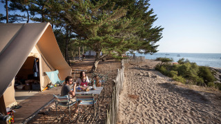 Familie nyder et måltid foran et Toile&Bois safari-telt ved kysten med udsigt over havet og træer.