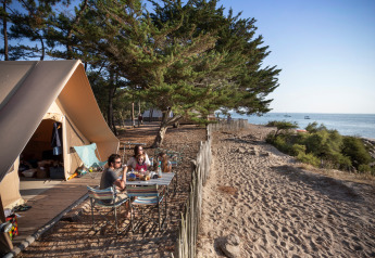 Familia disfruta de una comida frente a una tienda safari Toile&Bois junto a la costa, con vista al mar y árboles.