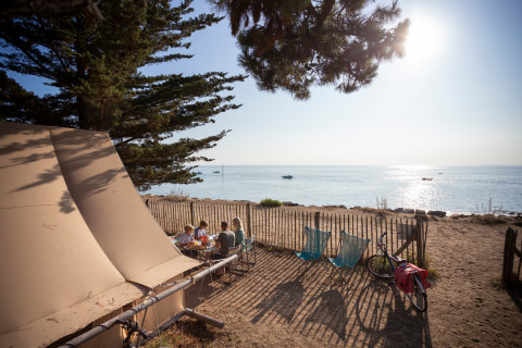 Familia comiendo frente a una tienda Toile&Bois en la costa de Huttopia Noirmoutier, Francia, con vista al mar.