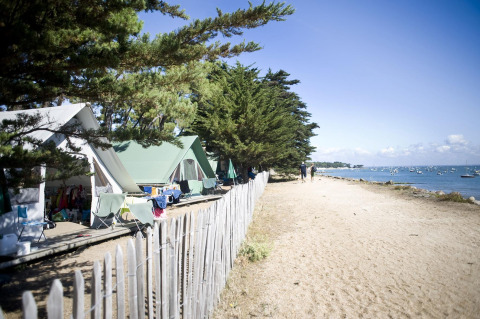 Tiendas tipo safari bajo árboles junto a la costa en Huttopia Noirmoutier, Francia, con vista al mar y barcos.