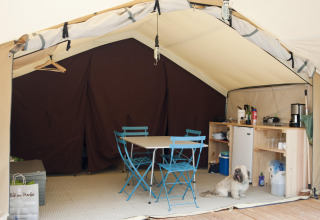 Interior de una tienda safari con sillas azules, mesa, cocina y un perro en Huttopia Calvados, Normandía.