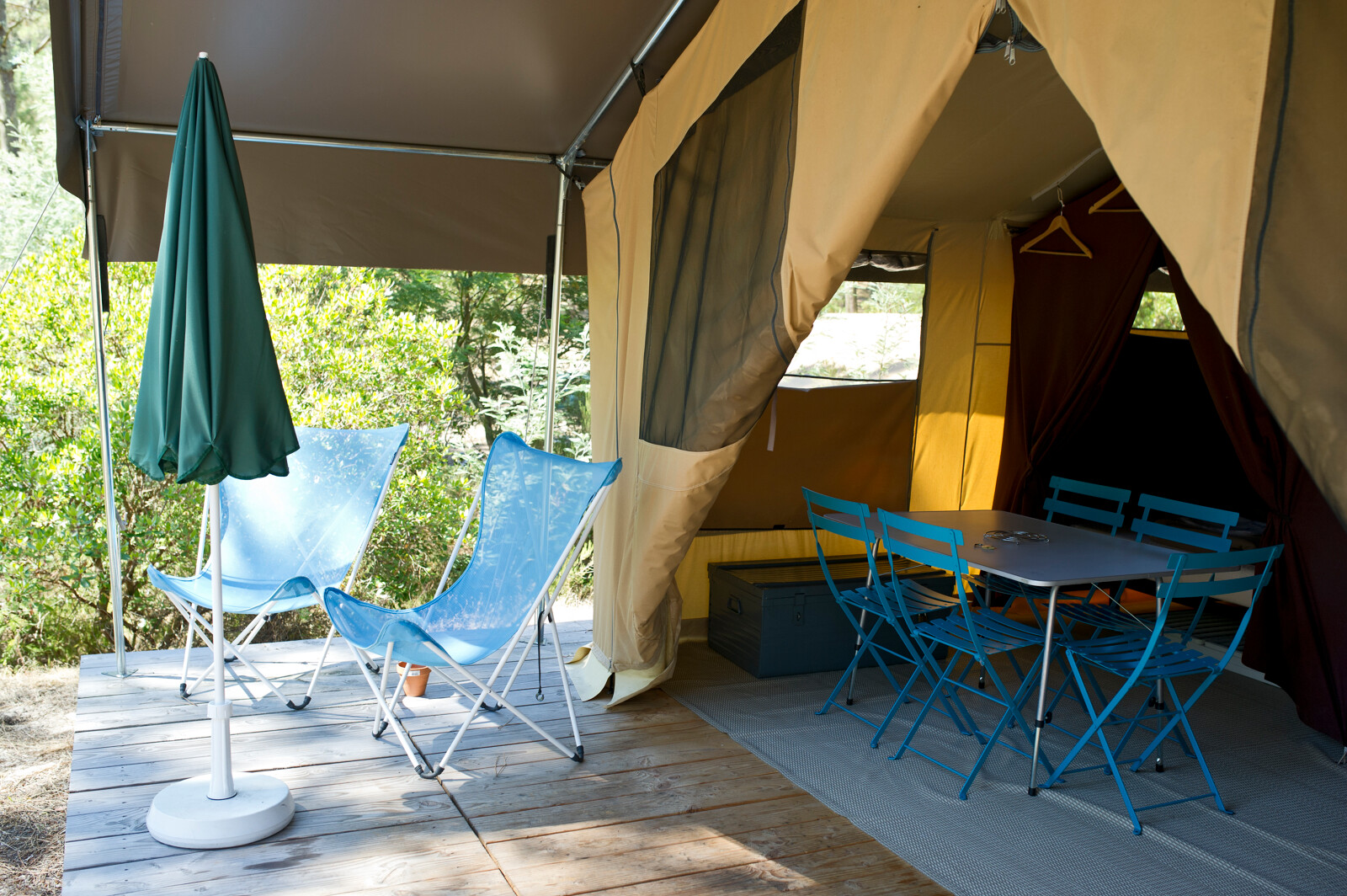 Safaritent met open ingang, twee blauwe stoelen, eettafel, stoelen en groene parasol op houten terras.