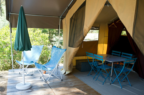 Safaritent met open ingang, twee blauwe stoelen, eettafel, stoelen en groene parasol op houten terras.