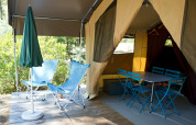 Safari-telt med åbent indgangsparti, to blå stole, bord, stole og en grøn parasol på en træterrasse.