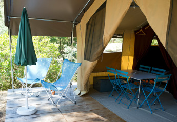 Tenda safari con ingresso aperto, due sedie blu, tavolo da pranzo, sedie e ombrellone verde su terrazza.