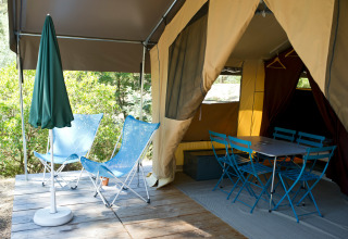 Tienda safari con entrada abierta, dos sillas azules, mesa comedor, sillas y sombrilla verde en terraza.