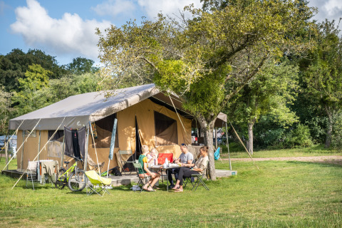 Familie sidder foran et safaritelte ved Huttopia Calvados - Normandie, Frankrig, omgivet af natur.