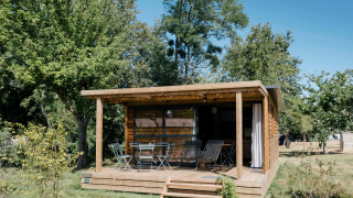 Chalet Evasion in legno con veranda coperta, sedie da giardino, immerso nel verde e sotto il sole.