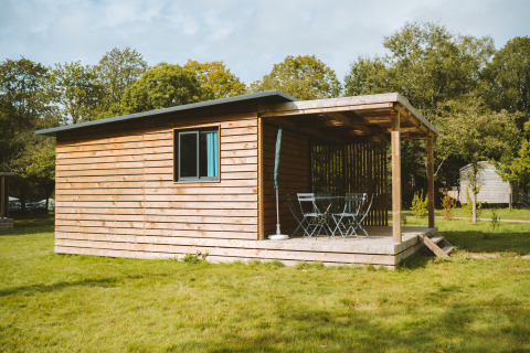Chalet de madera con terraza cubierta y muebles de exterior en césped en Huttopia Calvados - Normandie, Francia.