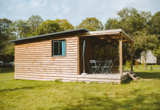 Chalet de madera con terraza cubierta y muebles de exterior en césped en Huttopia Calvados - Normandie, Francia.