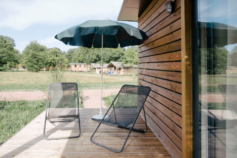 Udendørs terrasse ved Chalet Portland, Huttopia Calvados - Normandie, med stole, parasol og grøn udsigt.