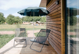 Udendørs terrasse ved Chalet Portland, Huttopia Calvados - Normandie, med stole, parasol og grøn udsigt.