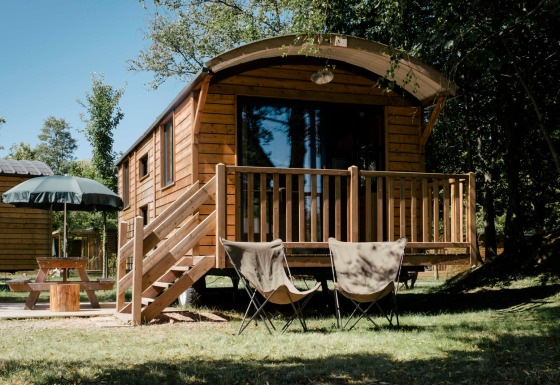 Cabaña de madera llamada Roulotte con porche, dos sillas y mesa de picnic en un entorno boscoso.