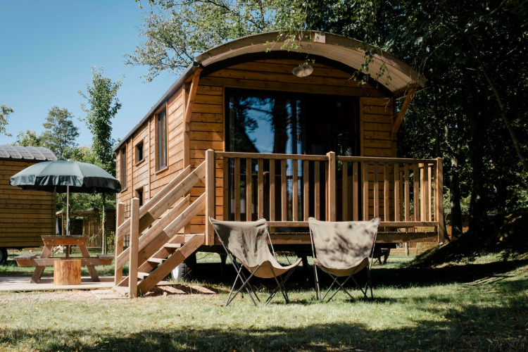 Cabaña de madera llamada Roulotte con porche, dos sillas y mesa de picnic en un entorno boscoso.