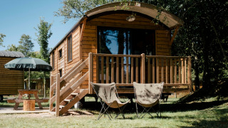 Cabaña de madera llamada Roulotte con porche, dos sillas y mesa de picnic en un entorno boscoso.