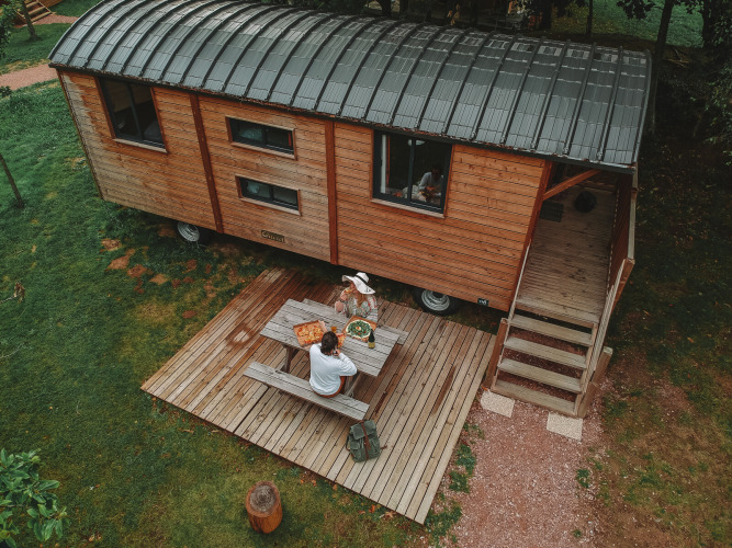 Vista aérea de una cabaña Roulotte de madera con terraza en Huttopia Calvados, Normandía, Francia, y dos personas comiendo.