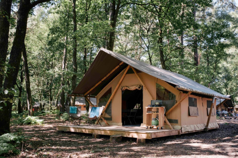 Tente safari Bungalow tent Trappeur poêle en forêt, terrasse en bois et chaises pliantes à l’extérieur.