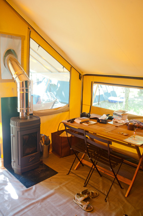 Interior de una tienda safari con estufa de leña y mesa, Bungalow tent Trappeur poêle en Huttopia Lac de Sillé, Francia.