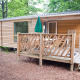 Træbeklædt mobilhome Cottage med veranda og udendørs spiseplads, omgivet af grønne træer.