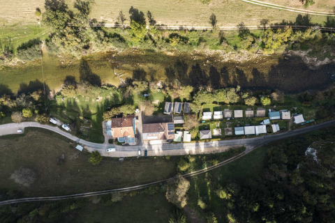 Luftfoto af feriepark med glamping-overnatning, campingvogne og grønne omgivelser tæt på en flod.
