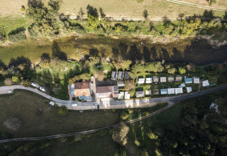 Vue aérienne d’un parc de vacances avec hébergements glamping, caravanes et verdure au bord d’une rivière.