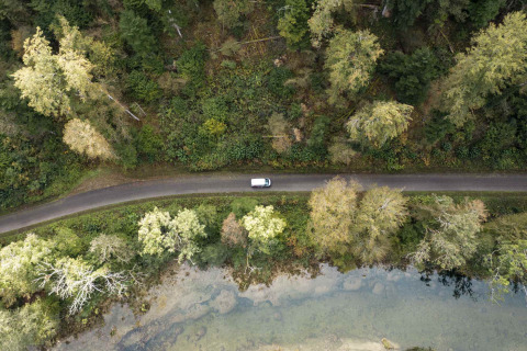 Vista aérea de un coche en una carretera junto a árboles y agua en un parque de glamping vacacional.