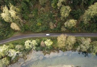 Vista aérea de un coche en una carretera junto a árboles y agua en un parque de glamping vacacional.
