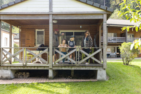 Una famiglia si trova sulla veranda di una casetta glamping in un villaggio vacanze circondato da verde e chalet.