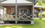 Une famille se tient sur la terrasse d'une cabane glamping dans un parc de vacances entouré de pelouse et chalets.