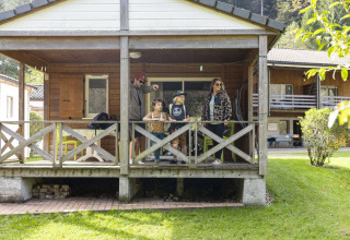 En familie står på verandaen af en glampinghytte i en feriepark med grønne omgivelser og træbygninger.