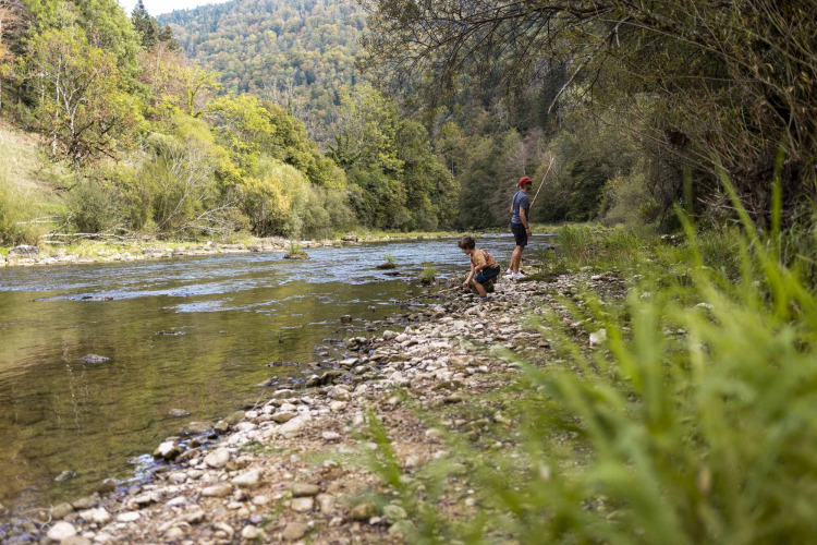 Deux personnes sur la berge d’une rivière arborée dans un parc de vacances avec hébergements glamping.