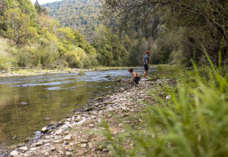 Two people on a rocky riverbank surrounded by trees, enjoying nature at a holiday park offering glamping.